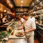 Chef prépare un plat devant des clients dans une cuisine ouverte chaleureuse